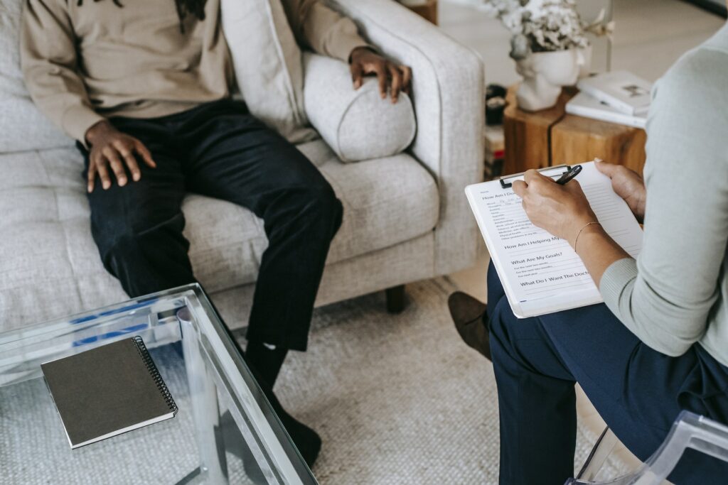 From above crop African American male in casual clothes sitting on comfy couch and answering professional female consultant questions in modern psychotherapy office